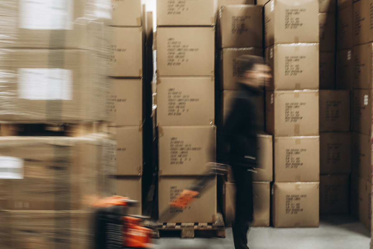 Blurred warehouse worker moving pallet handling equipment between tall stacks of cartons in a high-volume warehouse storage area.