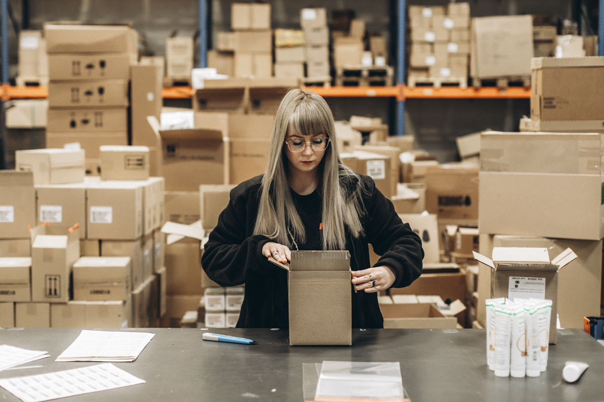 Warehouse employee packing products into a cardboard shipping box at a fulfillment workstation surrounded by inventory and packed cartons.