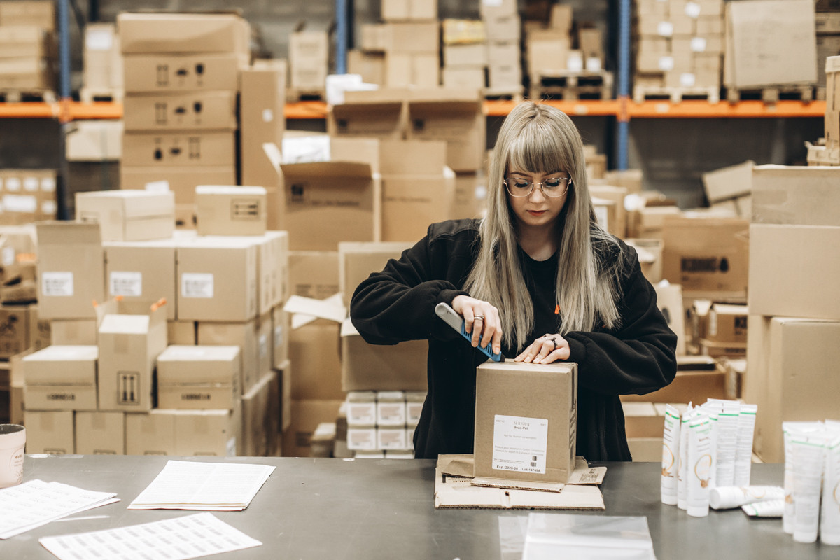 Warehouse employee packing products into a cardboard shipping box at a fulfillment workstation with inventory stock and packed cartons in the background.