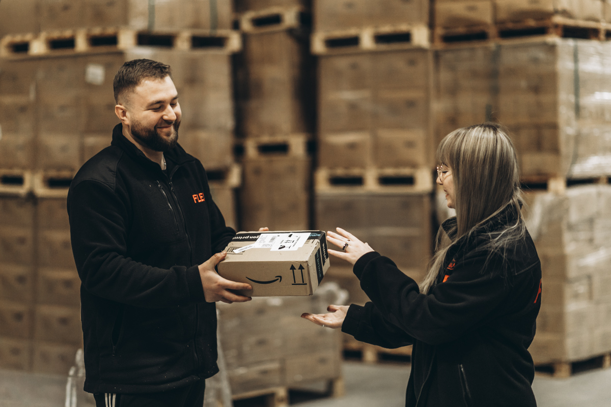 Two fulfillment warehouse employees exchanging a labeled parcel in front of palletized inventory and warehouse stock storage.
