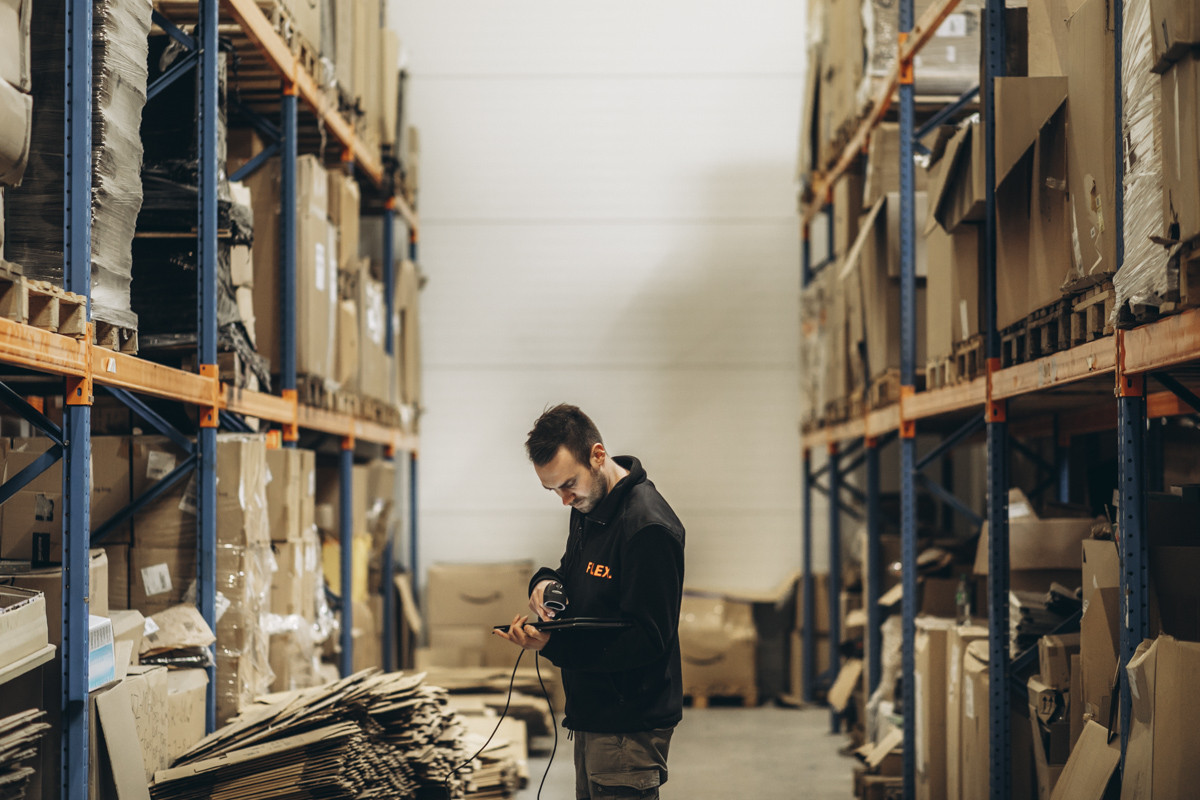 Warehouse employee using a handheld barcode scanner and tablet to check inventory in a storage aisle lined with pallet racking and stored cartons.