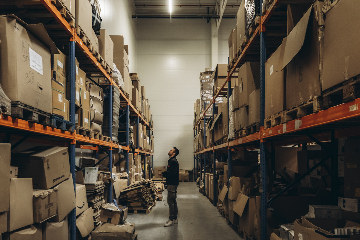 Warehouse employee conducting an inventory check in a storage aisle with pallet racking, cartons, and packaging materials stored on warehouse shelves.