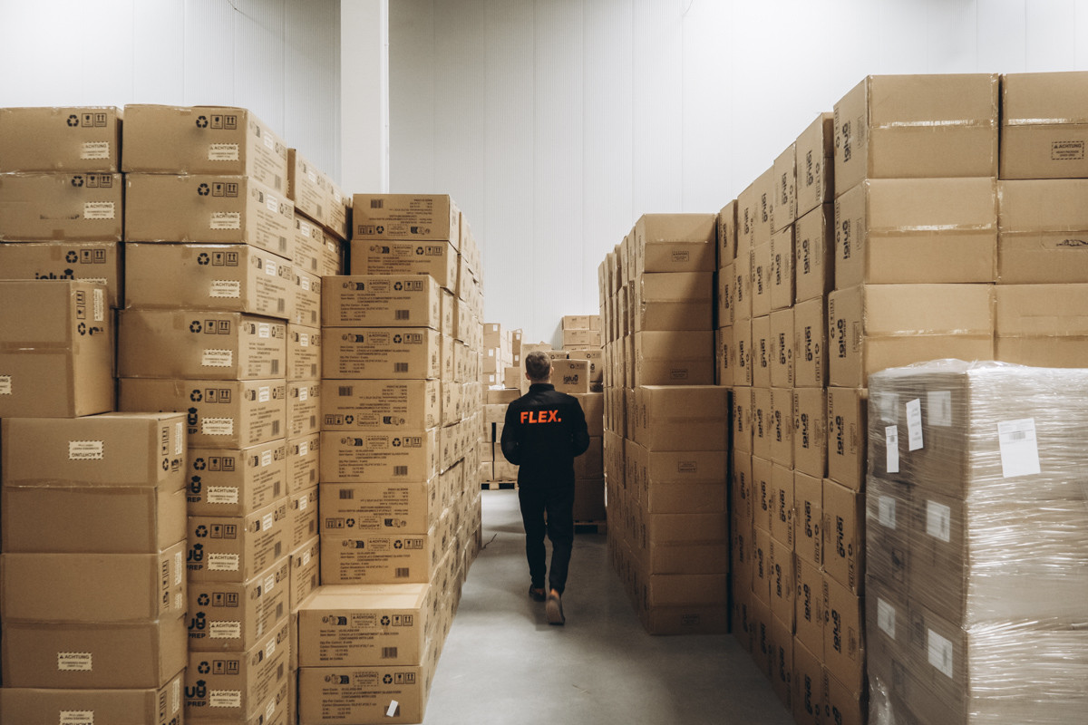 FLEX. warehouse employee walking through an aisle between tall stacks of cartons and palletized inventory in a large warehouse storage area.