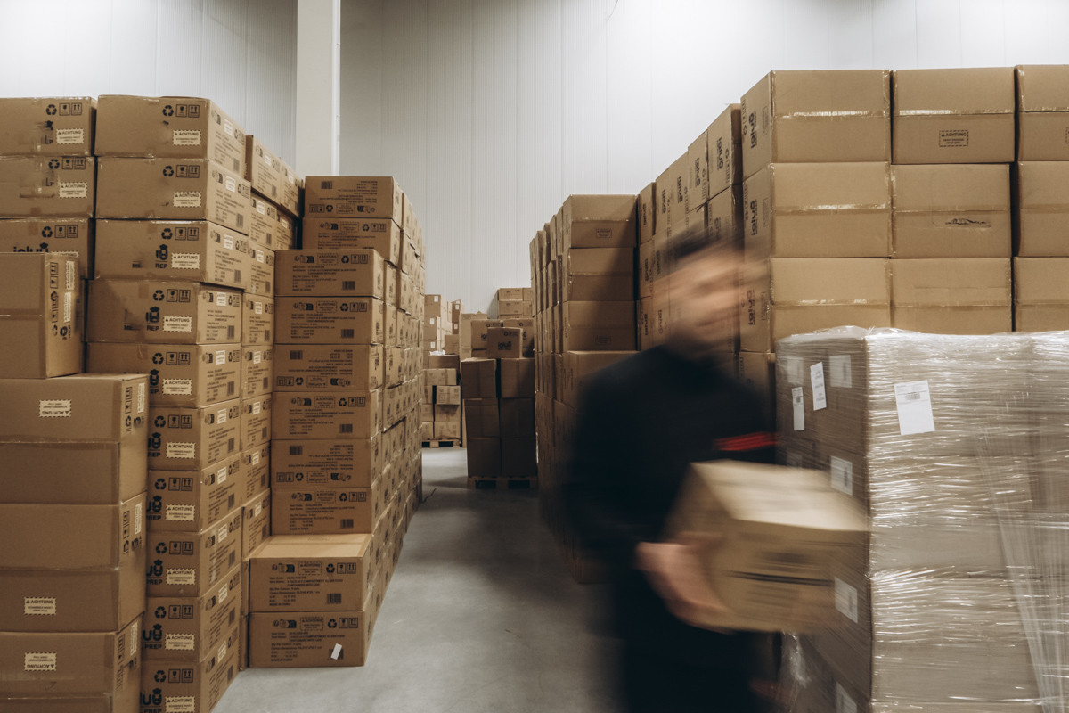 Blurred warehouse employee carrying a carton through an aisle between stacked inventory boxes and palletized stock in a warehouse storage area.