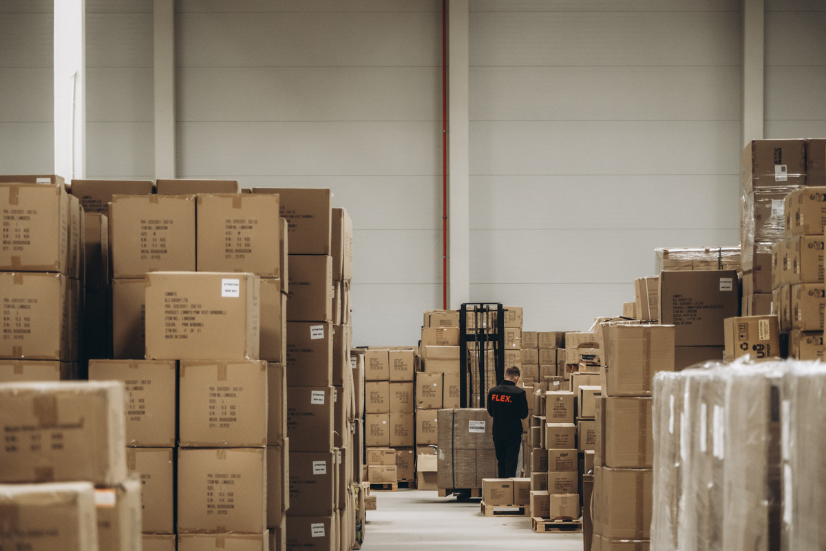Wide view of a warehouse storage area with stacked cartons, palletized inventory, and a FLEX. employee working among organized stock locations.