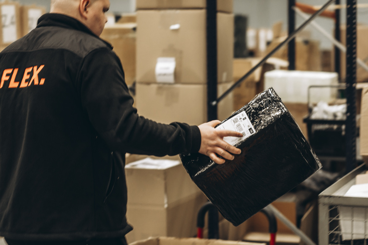 FLEX. warehouse worker inspecting a labeled parcel in a fulfillment warehouse surrounded by stacked cartons and inventory.
