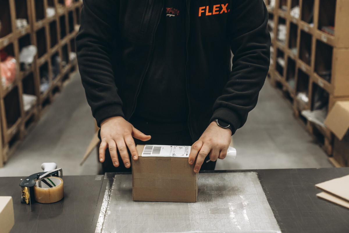 FLEX. warehouse worker applying a shipping label to a sealed cardboard parcel at a packing and dispatch workstation.